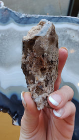 A hand holding a rough smokey quartz crystal with lodolite inclusions, showing earthy brown and beige tones against a blurred blue and white agate background.