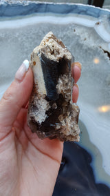 Hand holding a rough smokey quartz crystal with dark brown and black inclusions, surrounded by lighter beige and white mineral matrix, against a polished agate background.