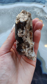 Hand holding a smokey quartz point with lodalite inclusions and beige mineral encrustations, against a background of a polished stone surface with blue and white banding.