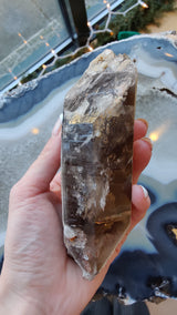 Hand holding a large smokey quartz crystal point with lodalite inclusions, featuring translucent brown and gray tones with a pointed top and natural rough base.
