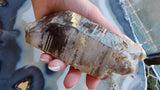 Hand holding a translucent smokey quartz crystal with visible lodalite inclusions and a rough, natural surface with varied brown and gray tones.