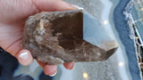 Hand holding a polished smoky quartz point with visible lodalite inclusions, showing a gradient from smoky brown to translucent gray with natural mineral textures