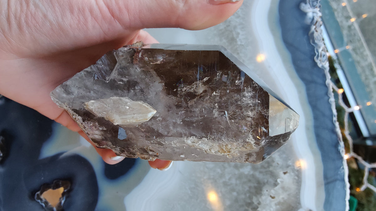 Person holding a pointed smoky quartz crystal containing white lodolite inclusions, shown over a polished agate slice with concentric blue and white bands.