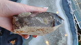 Hand holding a smokey quartz point with lodalite inclusions, showing translucent gray and beige tones with internal textures.