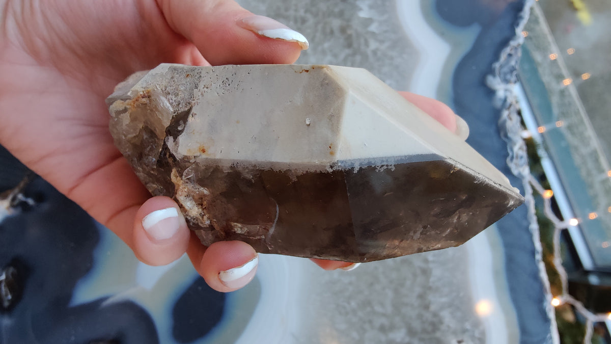 Hand holding a pointed smoky quartz crystal with lodolite inclusions, showing smooth and rough facets in shades of brown and beige.