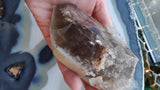 Hand holding a translucent smokey quartz crystal point with visible lodalite inclusions and natural rough edges, set against a blue and white agate surface with string lights in the background.