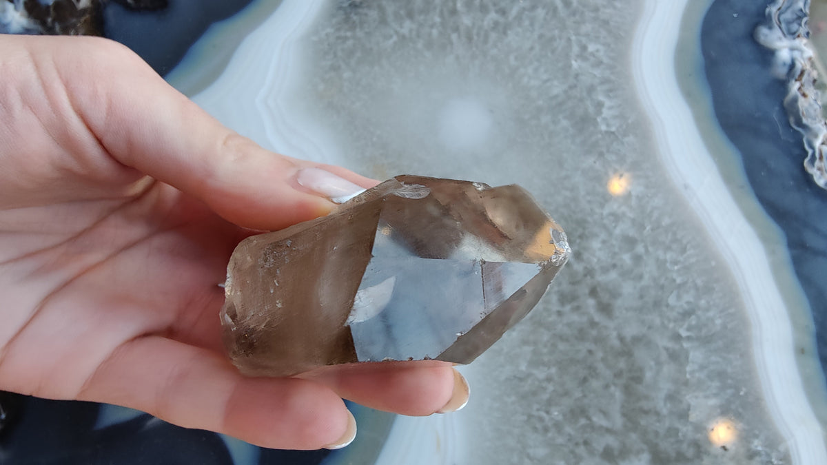 Hand holding a translucent smoky quartz crystal with visible internal lodolite inclusions, against a background of a gray and white banded agate slice.