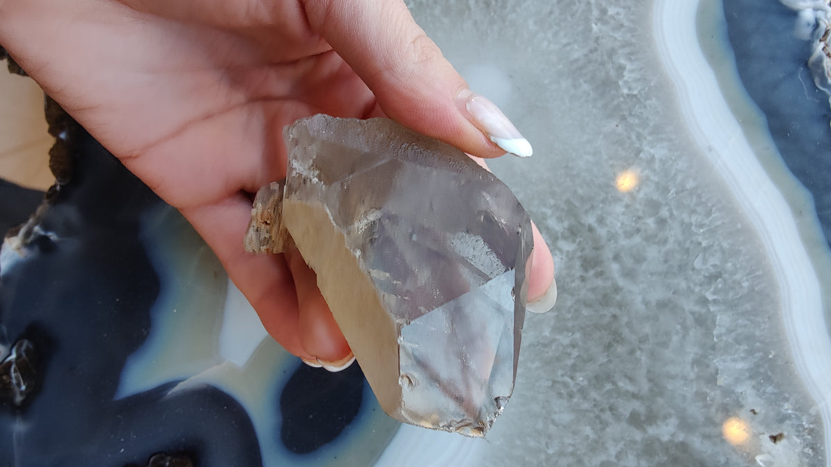 Hand holding a pointed smoky quartz crystal with visible lodolite inclusions, set against a background of textured gray and blue agate slices.