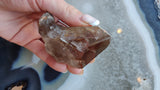 Translucent smoky quartz crystal with brown and gray inclusions, held by a hand with manicured nails, resting above a polished blue and white stone surface.