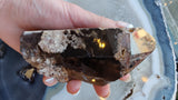 Hand holding a large smoky quartz point featuring dark translucent surfaces with white lodolite mineral inclusions inside, against a pale blue agate background.