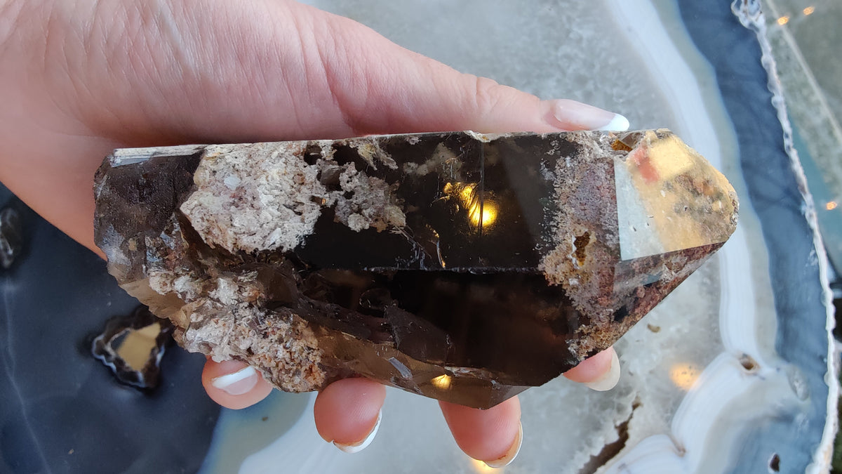 Hand holding a large smoky quartz point featuring dark translucent surfaces with white lodolite mineral inclusions inside, against a pale blue agate background.