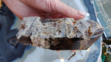 Hand holding a smoky quartz crystal with lodolite inclusions, displaying a rough, layered texture and a gradient from translucent gray to brown.