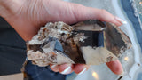 Hand holding a smoky quartz crystal with dark, translucent points and beige mineral inclusions, showing a rough, natural formation with angular edges.