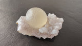 Translucent pale yellow fluorite sphere resting on a cluster of white quartz crystals with a rough, uneven surface, displayed on a dark textured background.