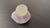 Translucent pale yellow fluorite sphere resting on a light purple quartz matrix with rough crystalline texture, shown on a dark brown surface.