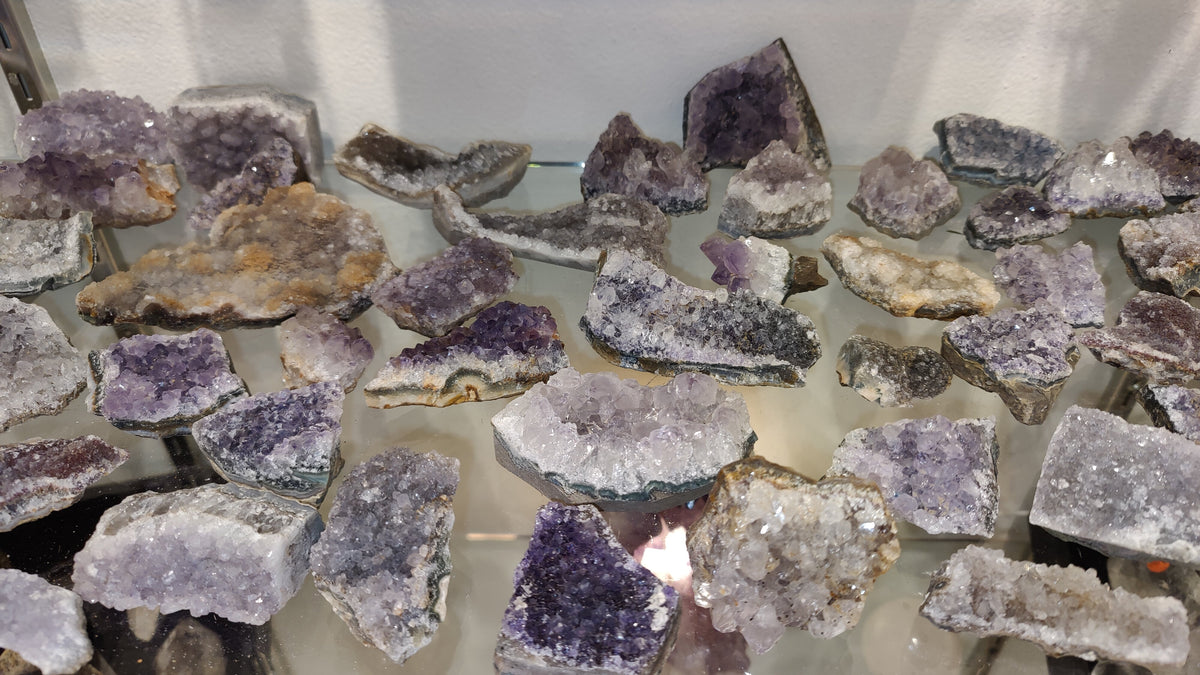 Collection of small Brazilian amethyst druze clusters displaying varying shades of purple and white crystalline formations on a reflective surface.