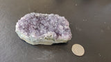 Small Brazilian amethyst druzy with dense light purple crystals on a grayish base, displayed next to a coin on a dark surface for scale.