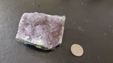 Small Brazilian amethyst druzy with light to medium purple crystals resting on a dark textured surface next to a quarter for size comparison.