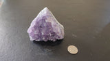 Small Brazilian amethyst druze with light to medium purple crystals on a white base, sitting on a dark gray textured surface beside a coin for scale.