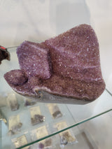 Close-up of a raw amethyst geode sculpture showing sparkling purple crystal formations within uneven rugged edges, displayed on a glass shelf with blurred labels underneath.