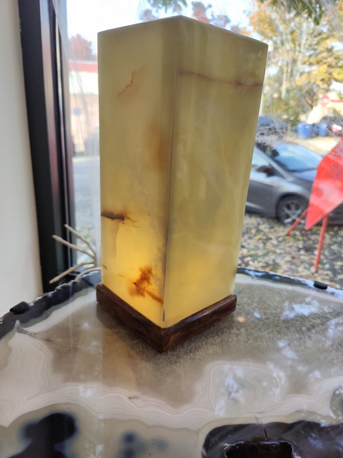 Tall square lamp with translucent green onyx sides showing natural brown veining, set on a dark wood base, illuminated indoors near a window with autumn foliage outside.