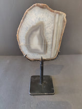Gray and white agate slice with concentric banding and crystalline texture, mounted vertically on a black metal stand against a neutral background.
