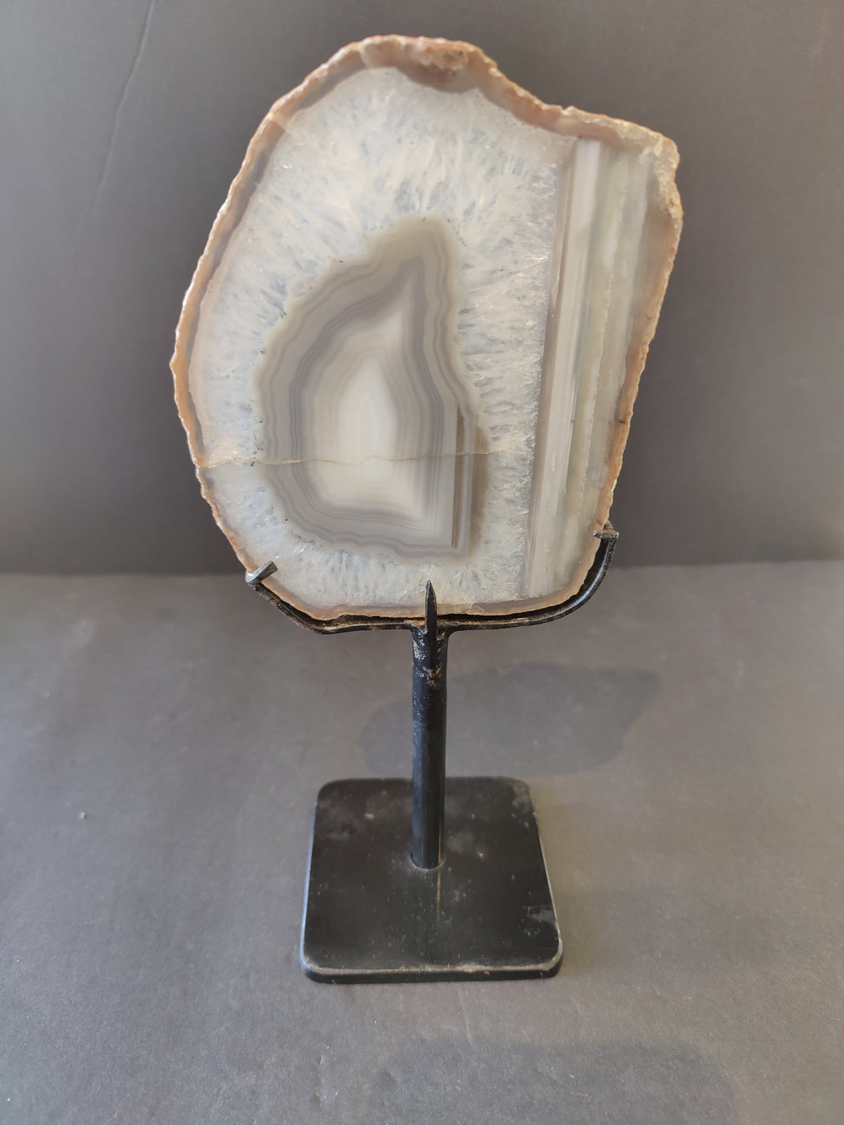 Gray and white agate slice with concentric banding and crystalline texture, mounted vertically on a black metal stand against a neutral background.