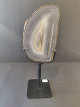 Vertical agate slice with concentric gray, white, and beige bands mounted on a black metal stand against a plain gray background.
