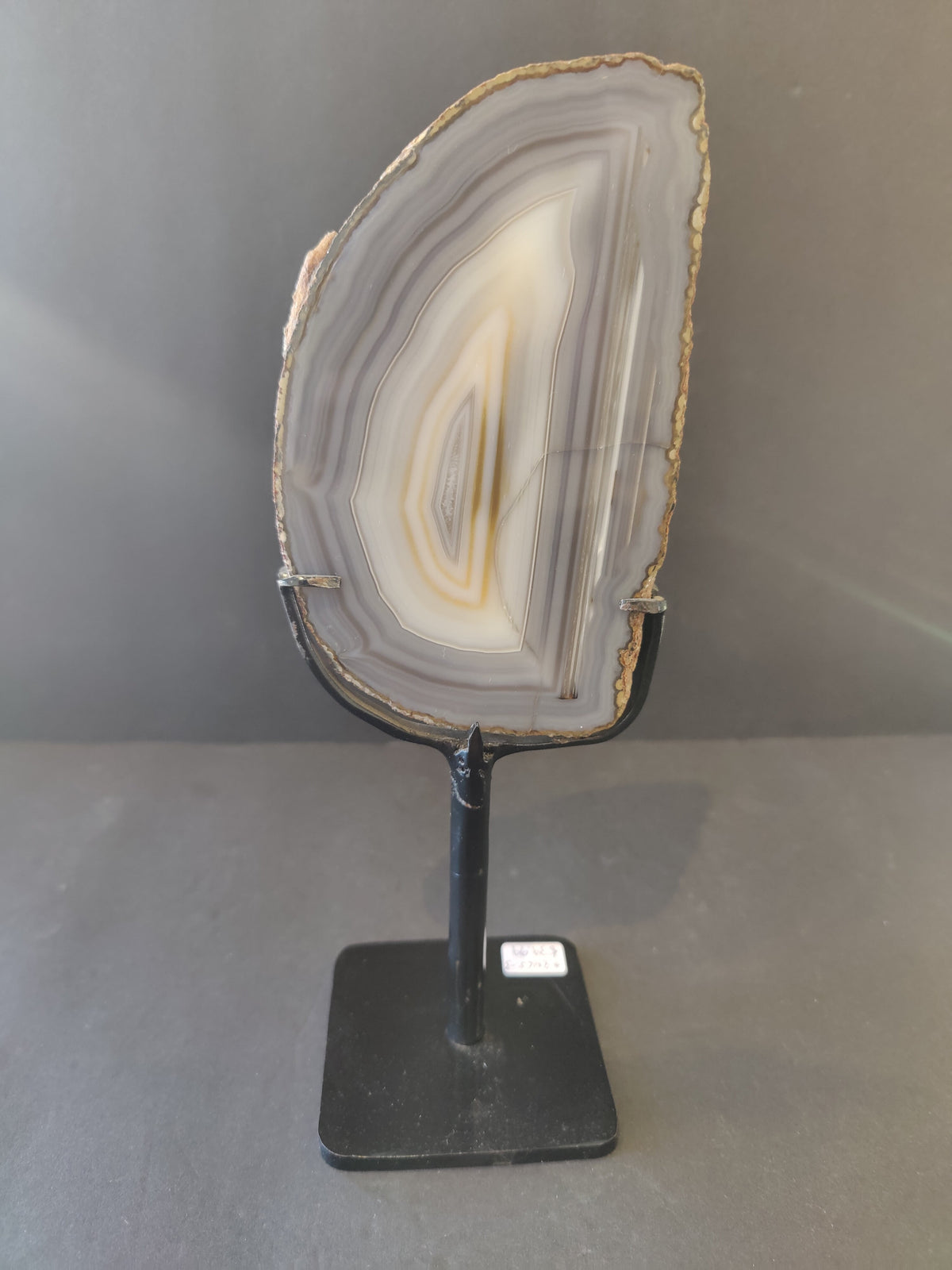 Vertical agate slice with concentric gray, white, and beige bands mounted on a black metal stand against a plain gray background.