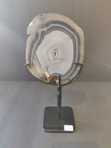 Translucent circular agate slice with concentric gray and white bands mounted vertically on a minimalist black metal display stand with a square base.