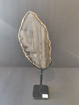 Vertical gray and beige agate slice with natural banding and rough edges mounted on a simple black metal stand against a gray background.