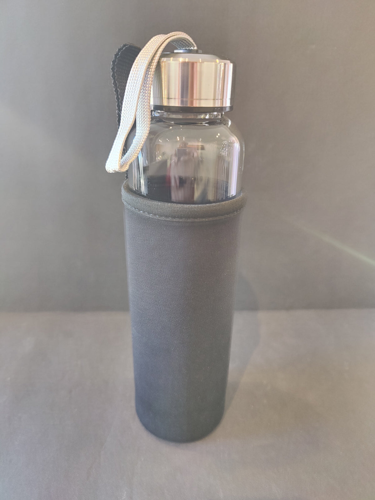 Clear glass water bottle with a silver metal screw cap and a white carrying strap, encased in a smooth dark gray insulating sleeve against a plain gray background.