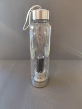 Clear glass water bottle with silver metal cap and base, containing a black crystal infused stainless steel rod inside, with a white carrying strap attached to the cap.
