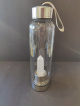 Transparent glass water bottle with silver metal cap and base, featuring a clear pointed crystal embedded inside the base and a beige carrying strap attached to the cap.