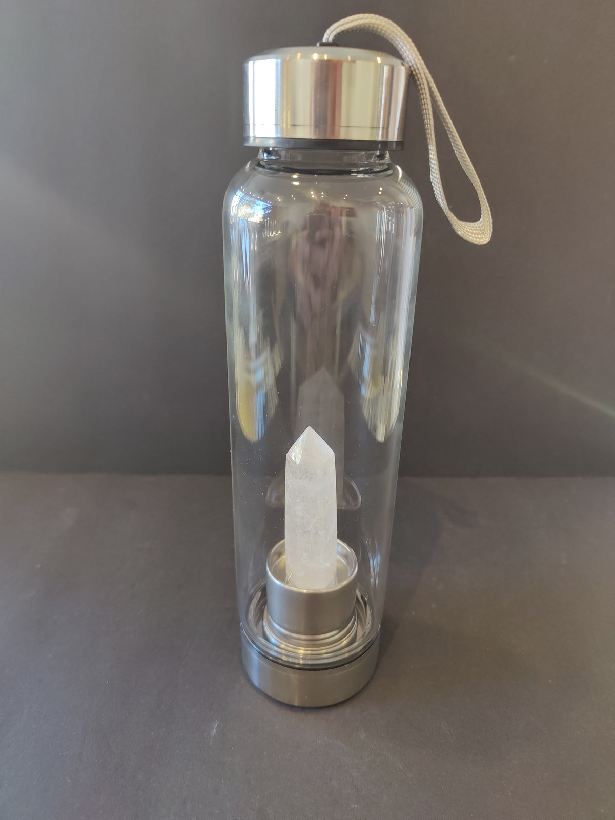 Transparent glass water bottle with silver metal cap and base, featuring a clear pointed crystal embedded inside the base and a beige carrying strap attached to the cap.