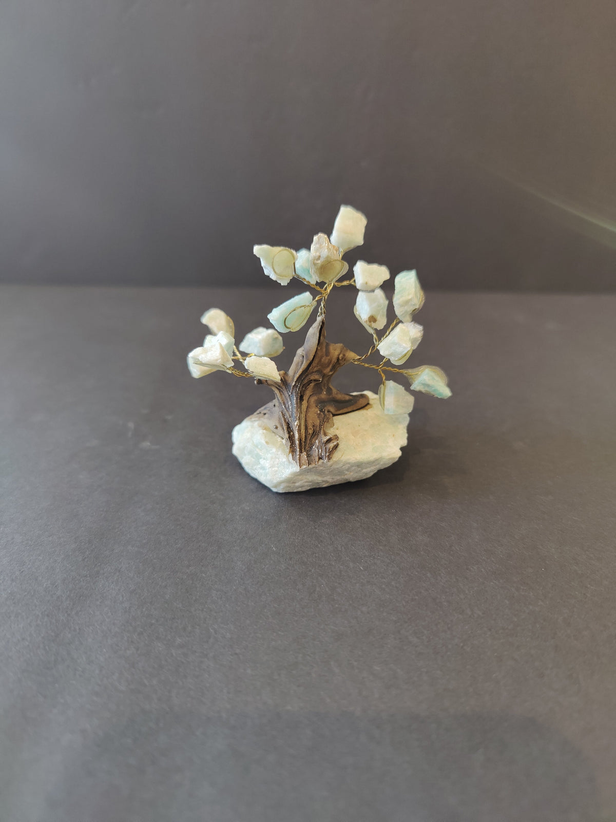 Small crystal bonsai tree sculpture with gold wire branches and white stone leaves mounted on a light-colored rough base, set against a dark gray background.