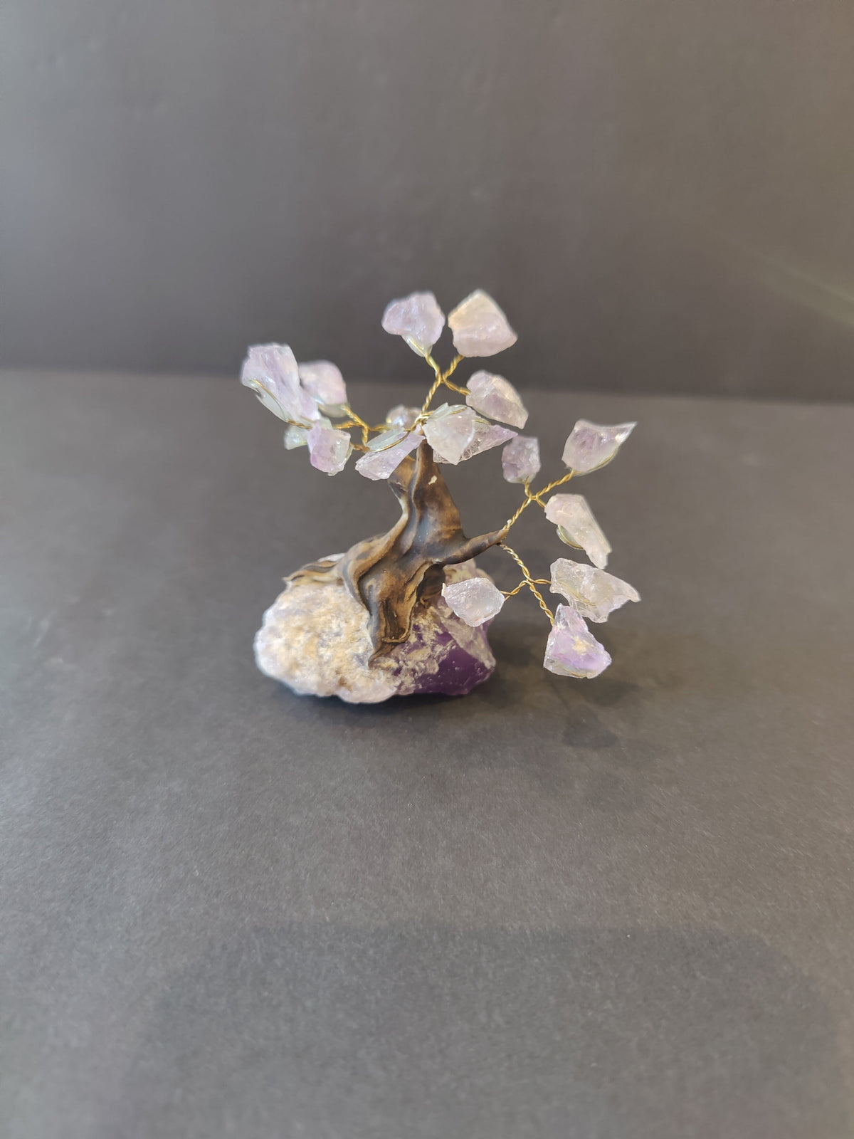 Small bonsai tree sculpture with twisted brown wire trunk and branches holding translucent purple crystal leaves on a rough purple and white rock base.