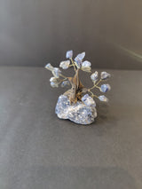 Small crystal bonsai tree sculpture with twisted brown wire trunk and branches holding translucent blue crystals, set on a rough blue-gray stone base against a gray background.