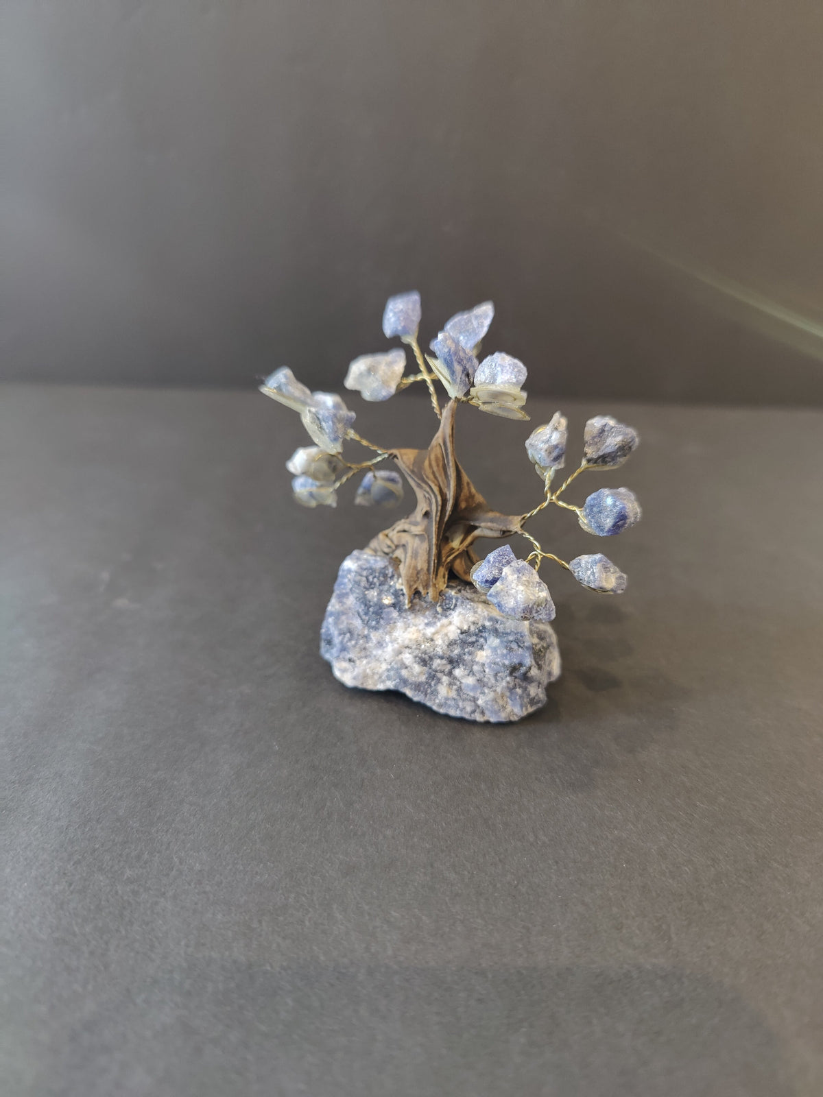 Small crystal bonsai tree sculpture with twisted brown wire trunk and branches holding translucent blue crystals, set on a rough blue-gray stone base against a gray background.
