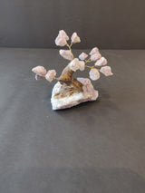 Small bonsai tree sculpture with pink crystal clusters attached to golden wire branches, mounted on a rough white and beige mineral base against a dark backdrop.