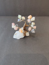 Miniature bonsai tree sculpture with a twisted brown trunk and multicolored crystal leaves mounted on a white rough crystal base, set against a gray backdrop.