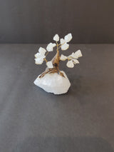 Small bonsai tree sculpture with white crystal leaves and twisted brown wire trunk, mounted on a rough white crystal base, photographed against a dark background.