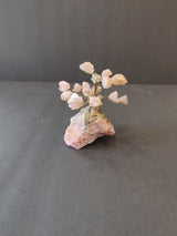 Small bonsai tree sculpture with pale pink crystal flower clusters and gold-colored wire branches, standing on a textured pink crystal rock base against a gray background.