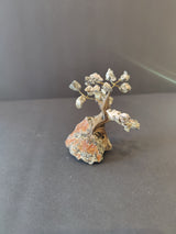 Small bonsai tree sculpture with a twisted brown trunk and branches holding crystal clusters, mounted on a rough, multicolored stone base against a gray backdrop.