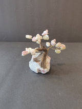 Small bonsai tree sculpture with twisted wooden trunk and pale pink and white crystal clusters as leaves, mounted on a rough white and pink stone base.