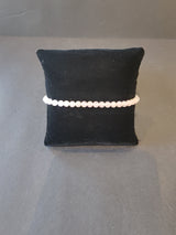 Delicate bracelet of small, light-toned, round beads displayed on a black fabric pillow against a gray surface.