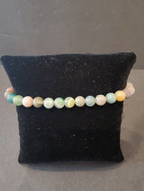 Single-strand bracelet with round 6mm beads in shades of green, cream, and light orange on a black fabric cushion.