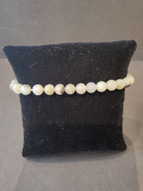 Close-up of a 6mm beaded stone bracelet with pale green and beige round beads displayed on a black velvet cushion against a dark background.