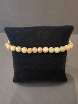 Circular beaded stone bracelet with 6mm round beads in mixed earthy tones of green, orange, and beige, displayed on a black velvet cushion.