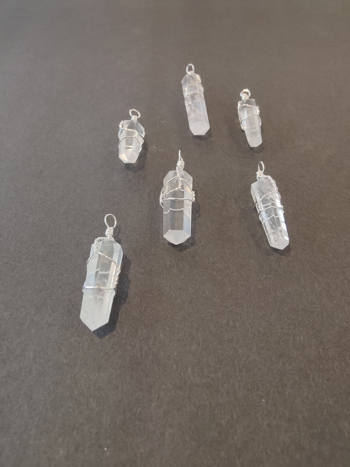 Six clear quartz crystal pendants wrapped in silver wire, arranged on a dark gray surface with varying shapes and wire styles.